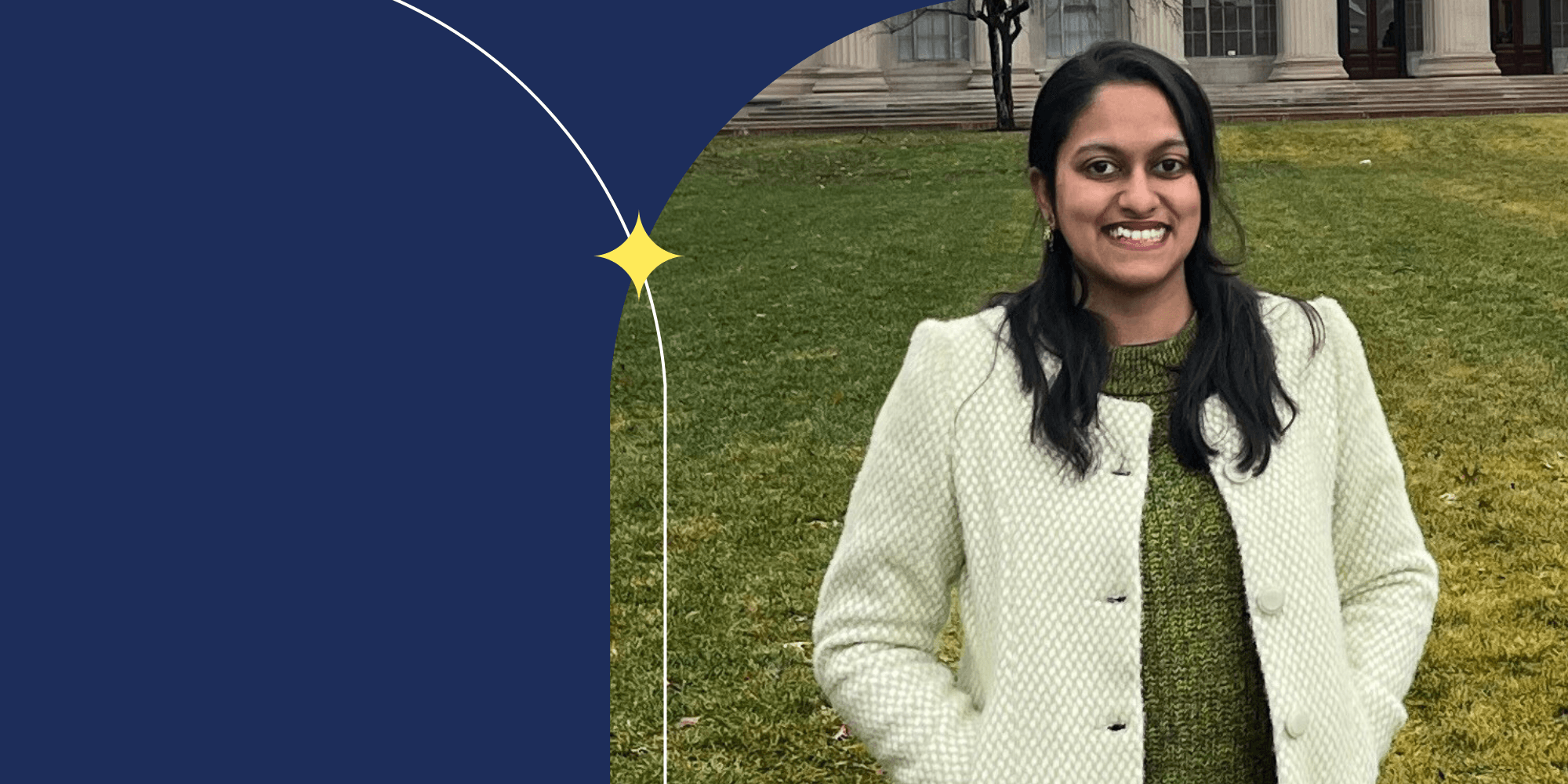 A smiling person standing in front of a building. They have long hair and are wearing a white jacket and a green shirt.